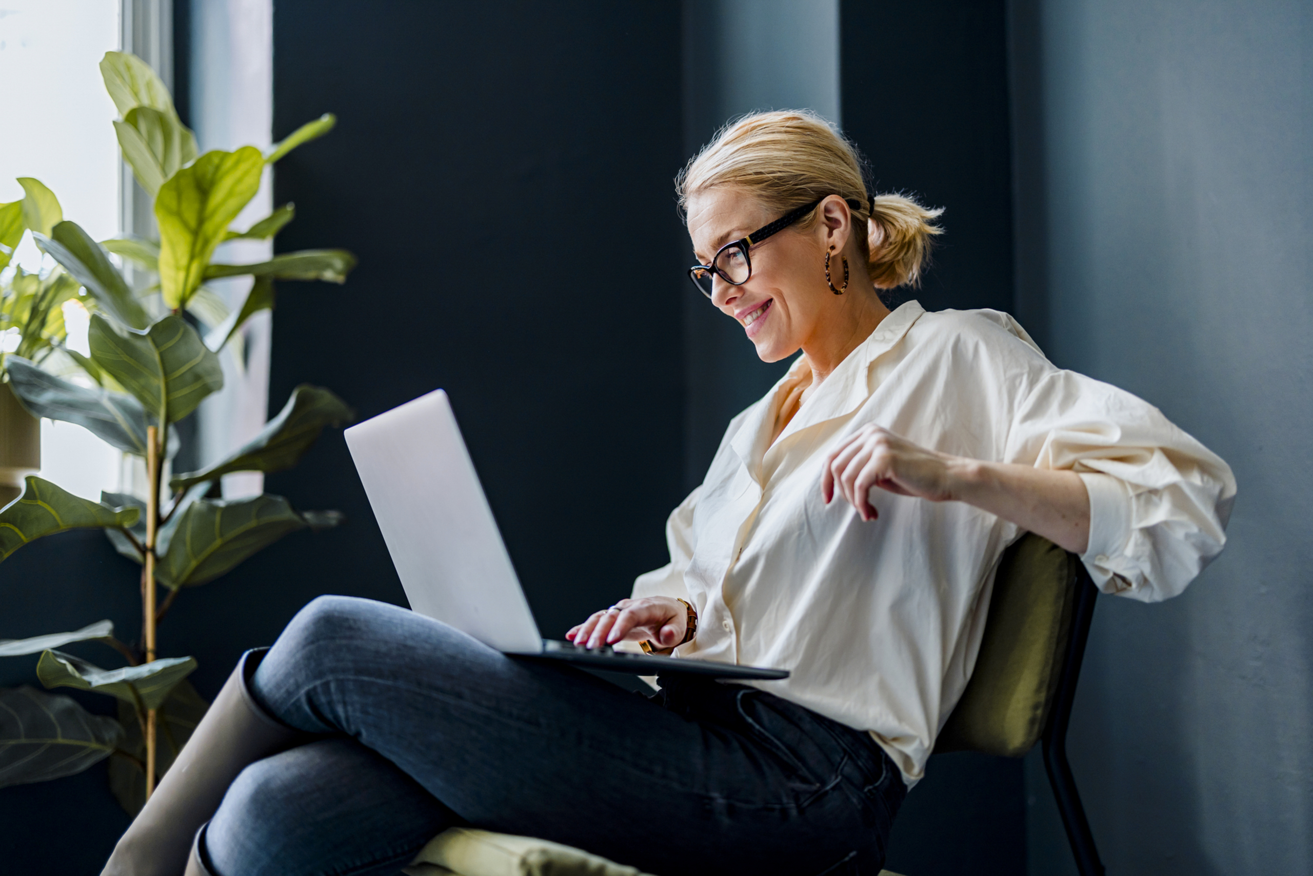 Cheerful smiling businesswoman sitting on a chair and typing business report on a laptop keyboard.