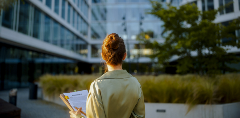 A woman looks at an office building whilst holding contract documents in her hand